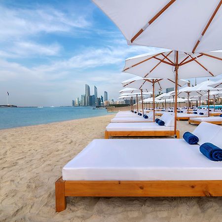 Radisson Blu Hotel & Resort, Abu Dhabi Corniche Extérieur photo The photo depicts a serene beach setting with several white lounge beds arranged neatly on the sandy shore. Each bed is paired with a white umbrella providing shade. In the background, a skyline of modern buildings can be seen along the coast, hintin