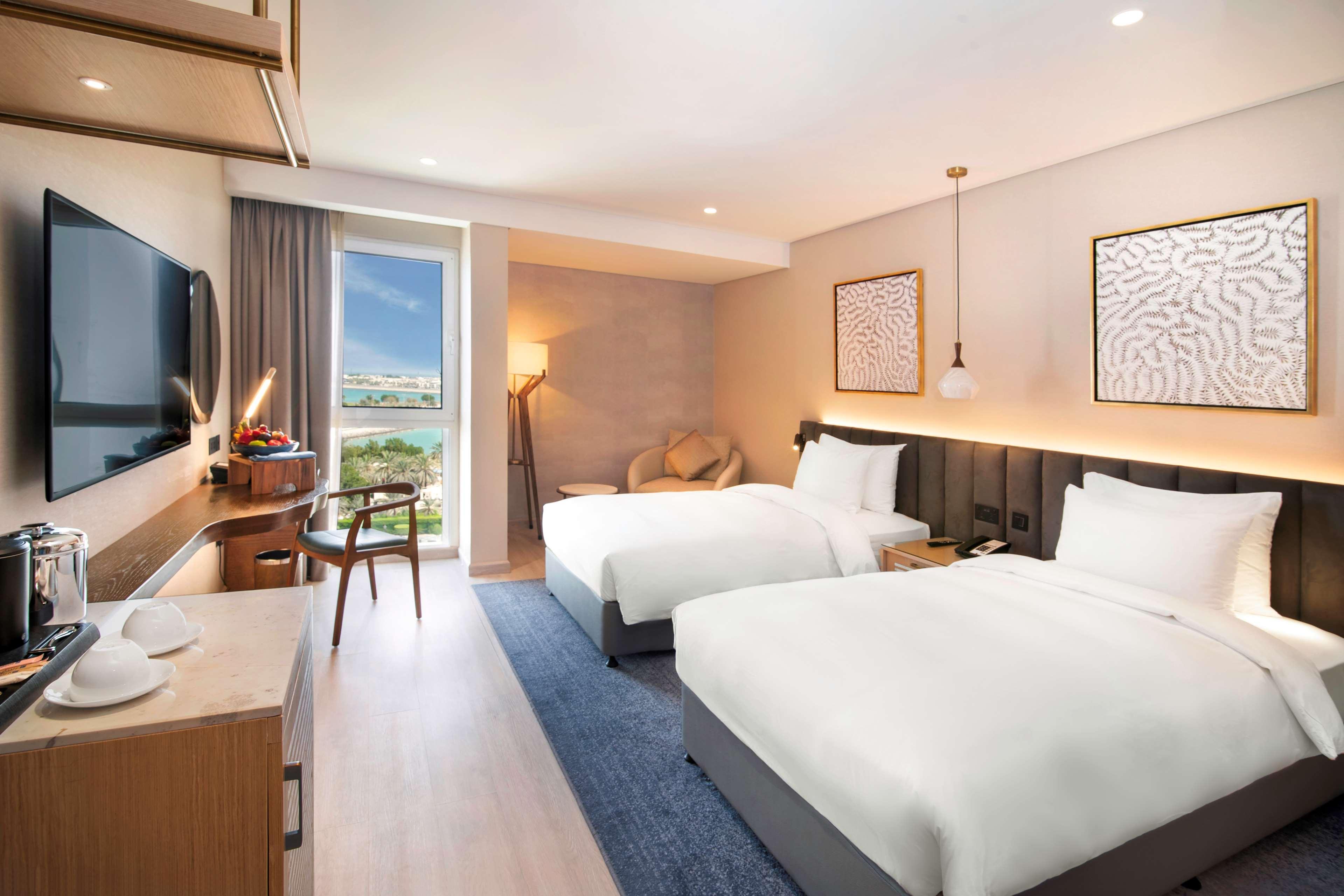 Radisson Blu Hotel & Resort, Abu Dhabi Corniche Extérieur photo The photo shows a modern hotel room featuring two beds with white bedding. There is a stylish desk and chair positioned near a large window that allows natural light to fill the space. The room has a contemporary design with muted colors and decorati