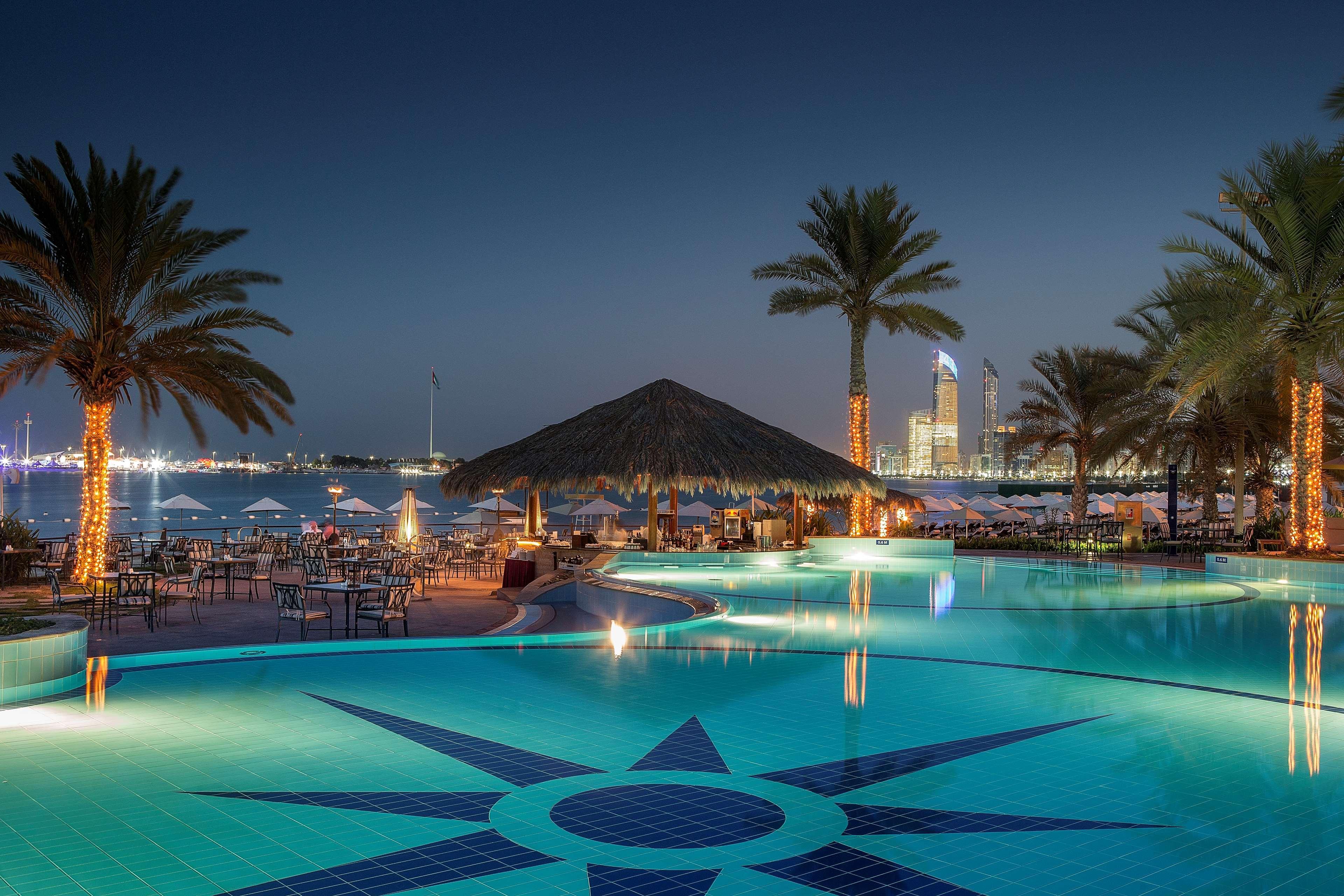 Radisson Blu Hotel & Resort, Abu Dhabi Corniche Extérieur photo The photo shows a picturesque outdoor pool area at night. The pool features a decorative tile pattern resembling a star at its center. Surrounding the pool are tall palm trees, some illuminated by soft lighting. In the background, there is a beachsid