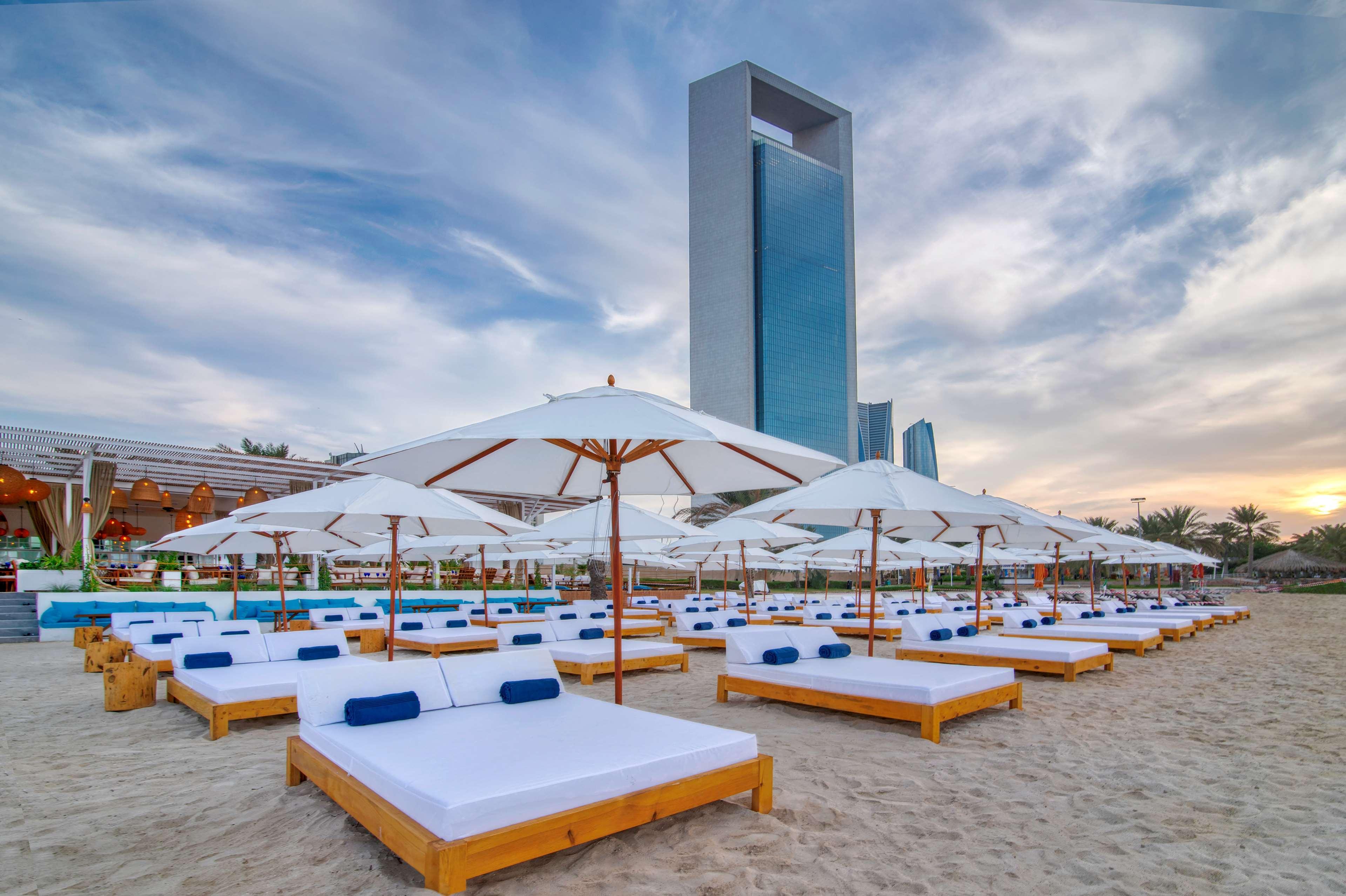 Radisson Blu Hotel & Resort, Abu Dhabi Corniche Extérieur photo The photo depicts a beach scene with a number of wooden sunbeds arranged on the sandy shore. Each sunbed is equipped with white cushions and blue pillows. Large white umbrellas are positioned above the sunbeds, providing shade. In the background, the