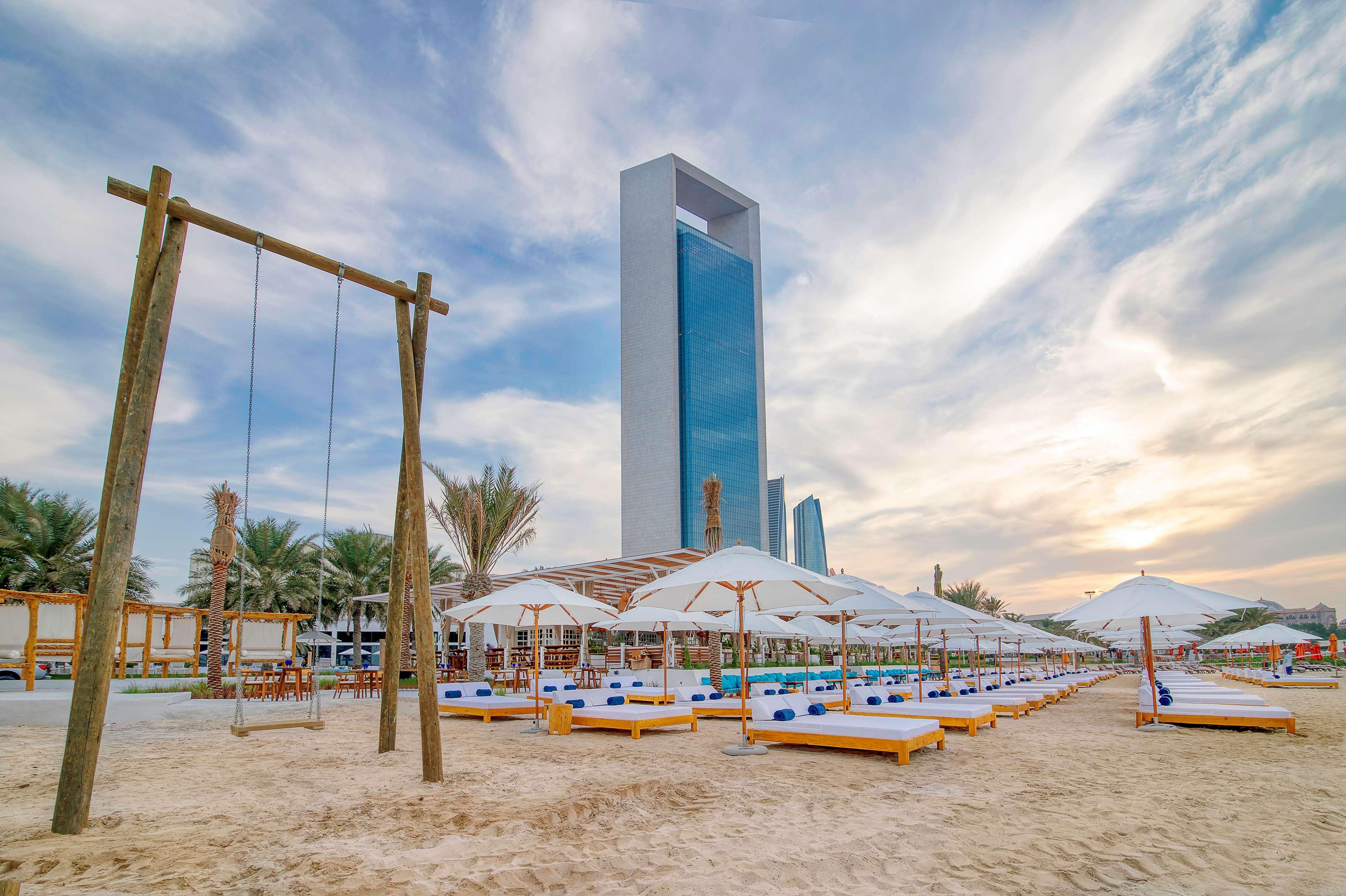 Radisson Blu Hotel & Resort, Abu Dhabi Corniche Extérieur photo The photo shows a beach scene with several sun loungers arranged under white umbrellas, set against a backdrop of a tall, modern building. There are palm trees in the area, providing a tropical ambiance. On one side, there's a wooden swing set, addin
