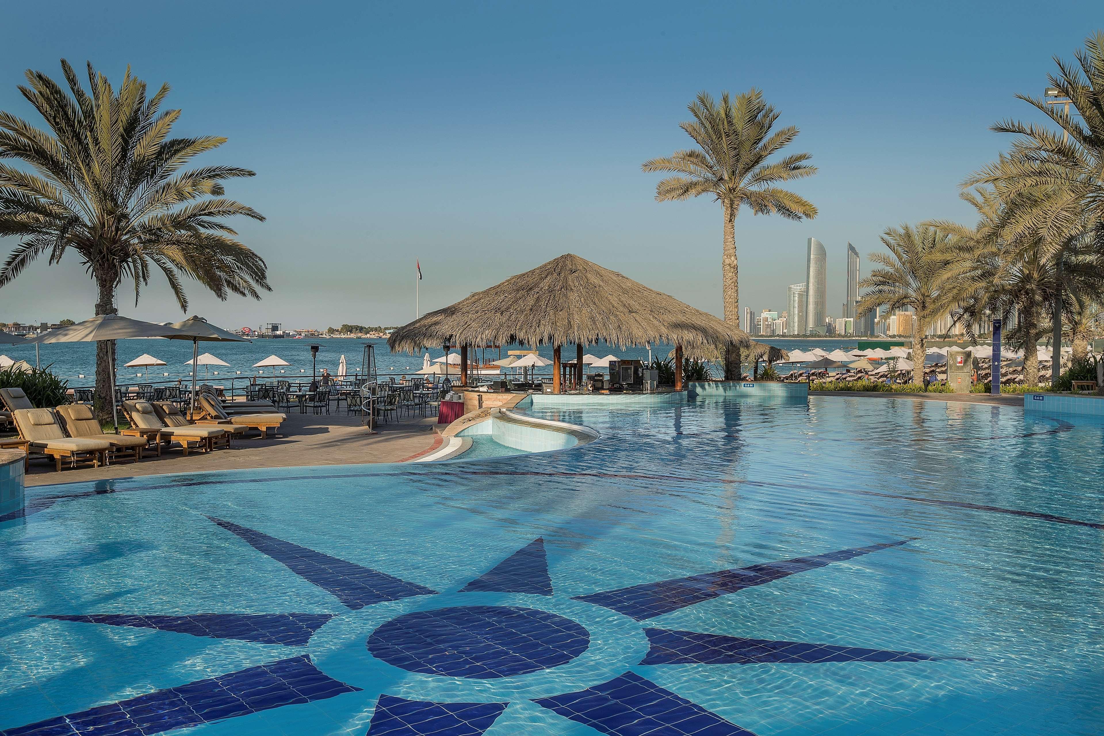Radisson Blu Hotel & Resort, Abu Dhabi Corniche Extérieur photo The photo shows a panoramic view of a luxurious resort pool area. In the foreground, there is a large blue swimming pool with a decorative star pattern on the bottom. Surrounding the pool are several lounge chairs, and there are palm trees providing 