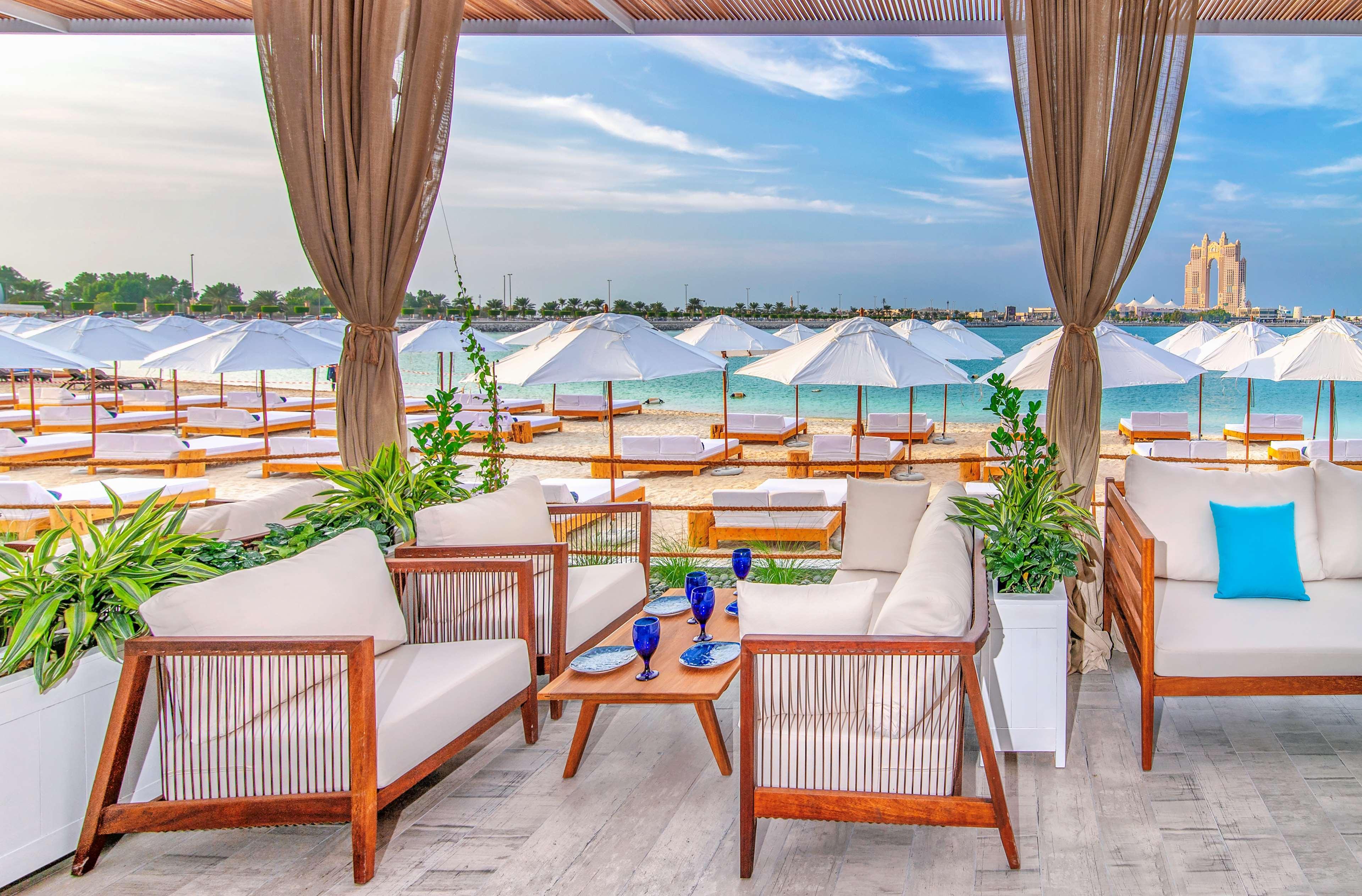 Radisson Blu Hotel & Resort, Abu Dhabi Corniche Extérieur photo The photo shows a stylish outdoor seating area at a beach or resort. There are comfortable lounge sofas arranged around a wooden coffee table, accompanied by decorative plants. In the background, a sandy beach is visible, lined with white umbrellas a