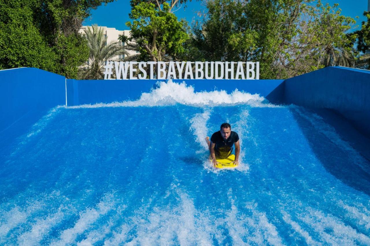 Radisson Blu Hotel & Resort, Abu Dhabi Corniche Extérieur photo The photo shows a person riding a bodyboard on a wave simulator, creating artificial waves. The background features a bright blue wall with the hashtag #WESTBAYABUDHABI displayed prominently. Surrounding the wave pool, there are trees and greenery, s