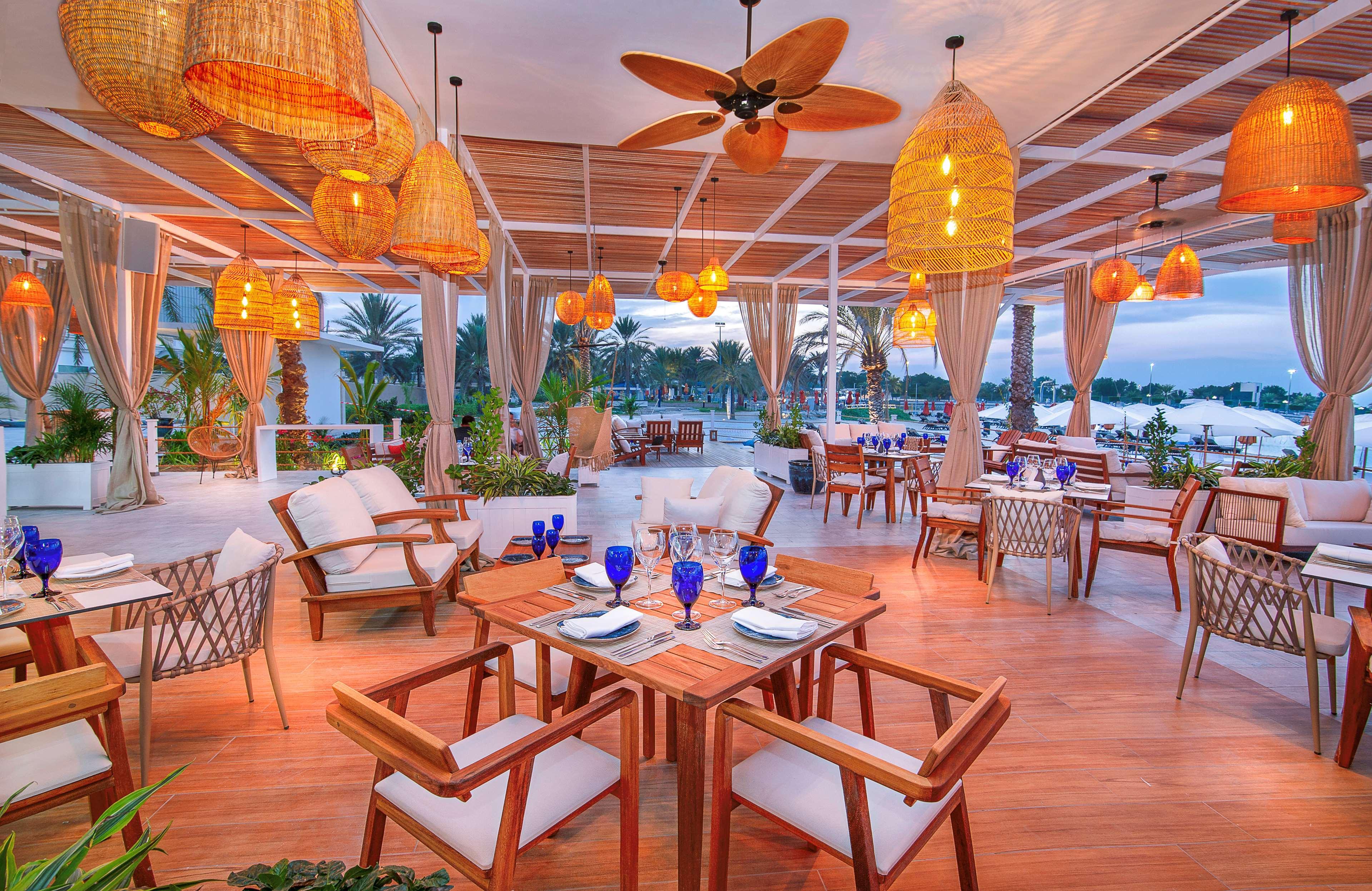 Radisson Blu Hotel & Resort, Abu Dhabi Corniche Extérieur photo The photo depicts an elegant dining area, likely in a restaurant. The space features wooden tables and comfortable chairs arranged neatly. There are multiple blue glasses set on the tables, suggesting a readiness for dining. The lighting is warm, pro