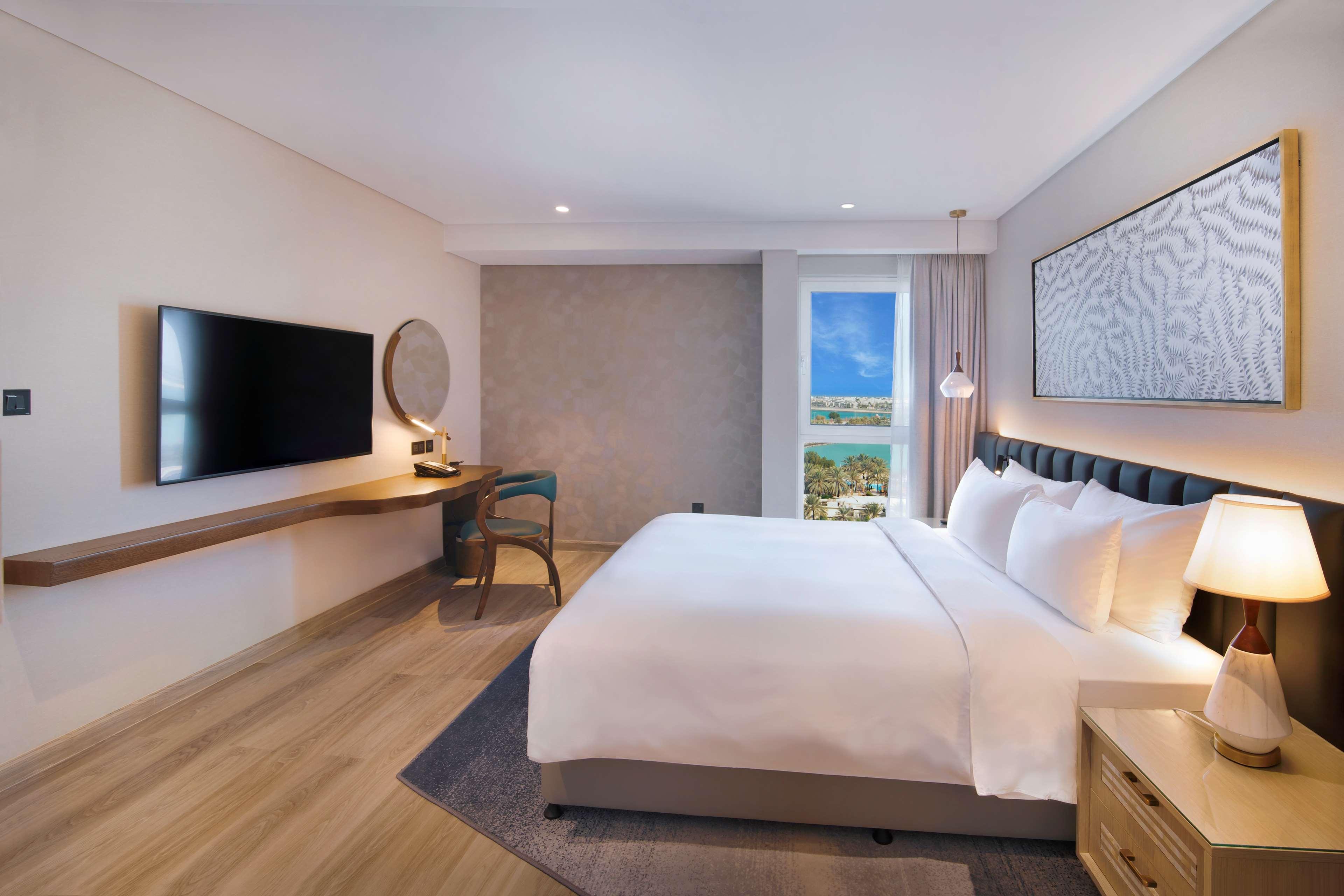 Radisson Blu Hotel & Resort, Abu Dhabi Corniche Extérieur photo The photo shows a modern hotel room with a clean and minimalistic design. It features a large bed with white bedding, complemented by bedside tables on either side with lamps. There's a wall-mounted television opposite the bed, and a desk with a chai