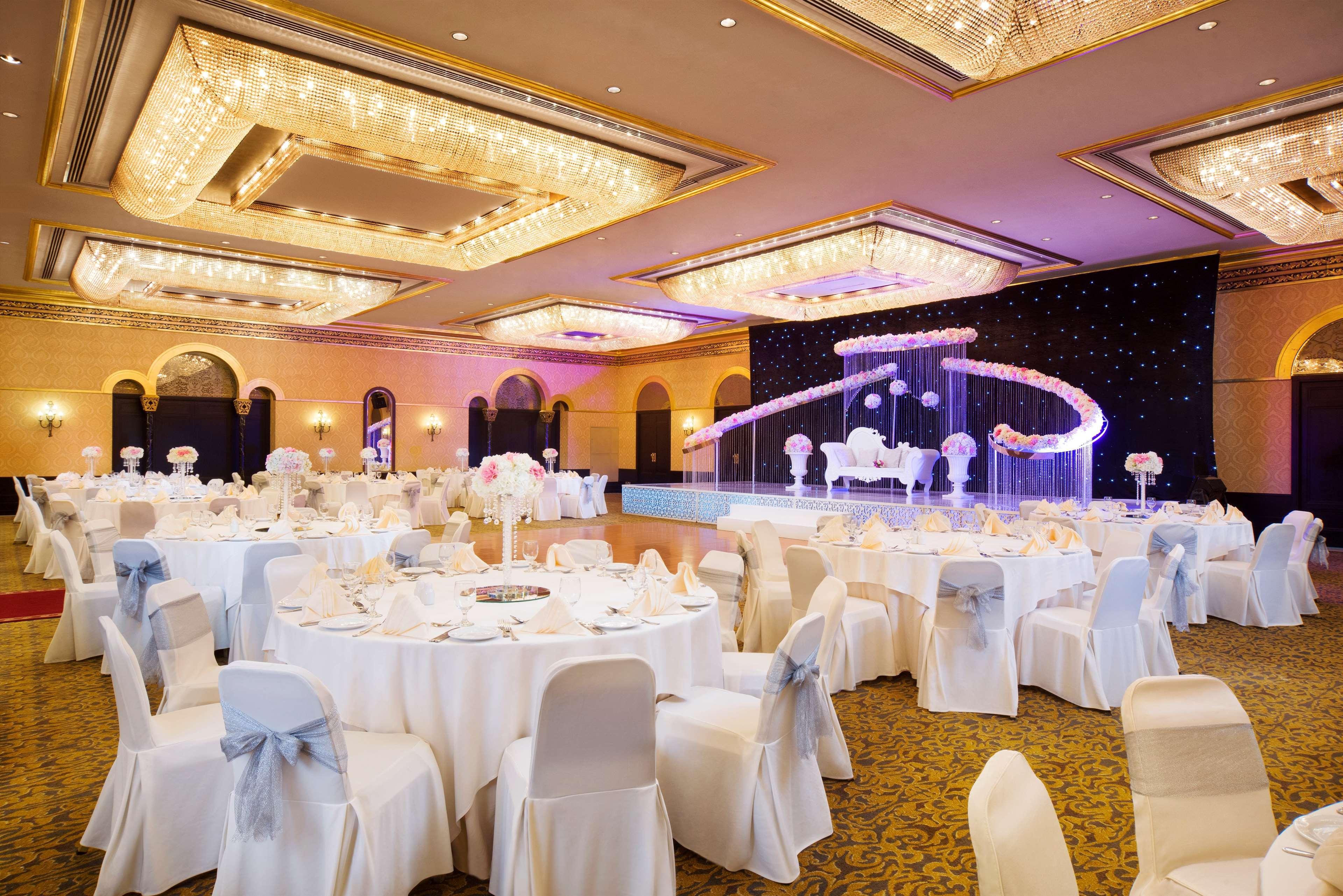 Radisson Blu Hotel & Resort, Abu Dhabi Corniche Extérieur photo The image shows an elegantly decorated banquet or event hall. There are several round tables covered with white tablecloths, each set with plates, cutlery, and folded napkins. The chairs are covered in white fabric with light blue sashes tied around 