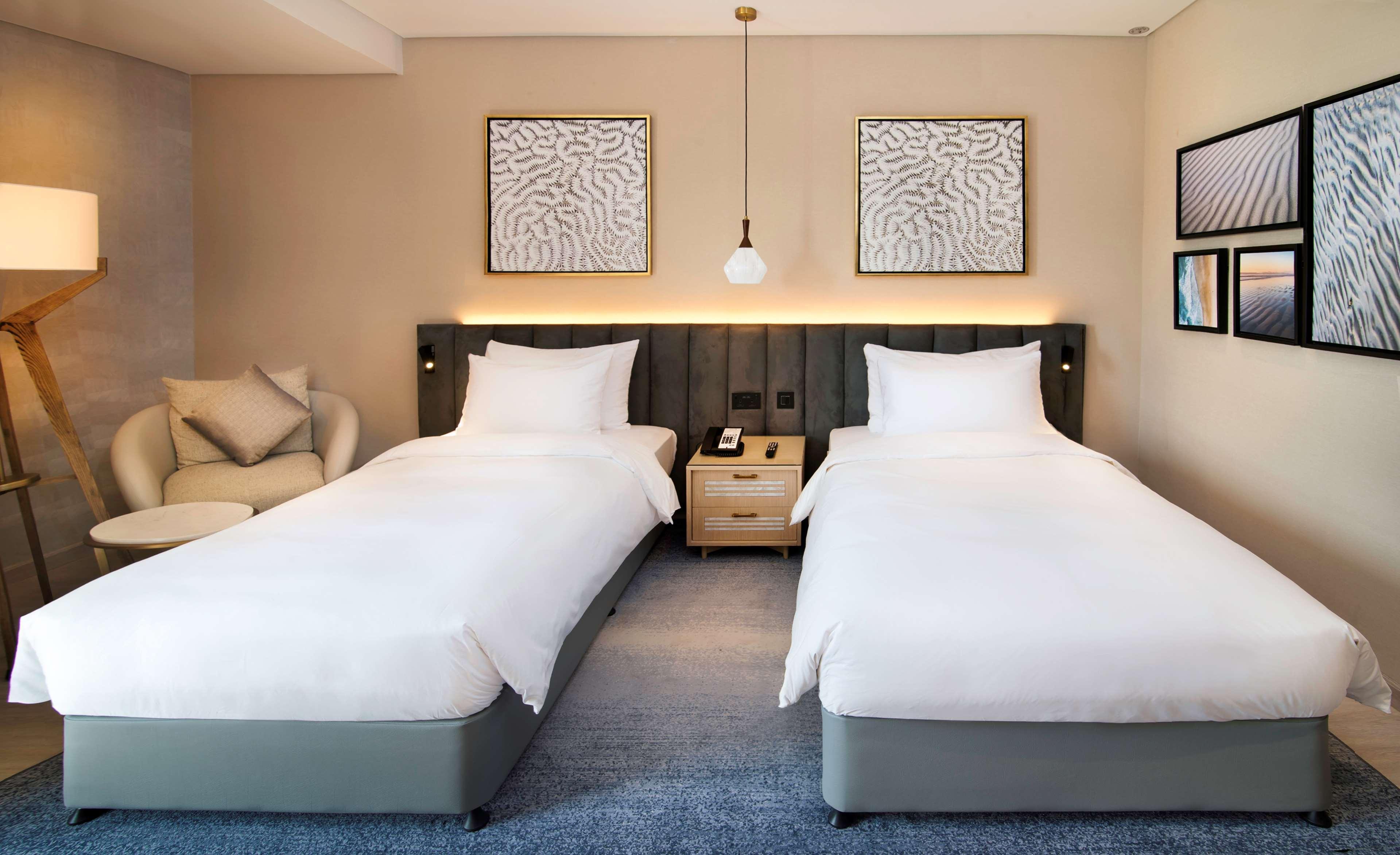 Radisson Blu Hotel & Resort, Abu Dhabi Corniche Extérieur photo The photo shows a modern hotel room with two single beds, each dressed in white bedding. The beds are positioned against a headboard with a gray color scheme. Above each bed, there are two framed artworks on the wall. A pendant light hangs from the c