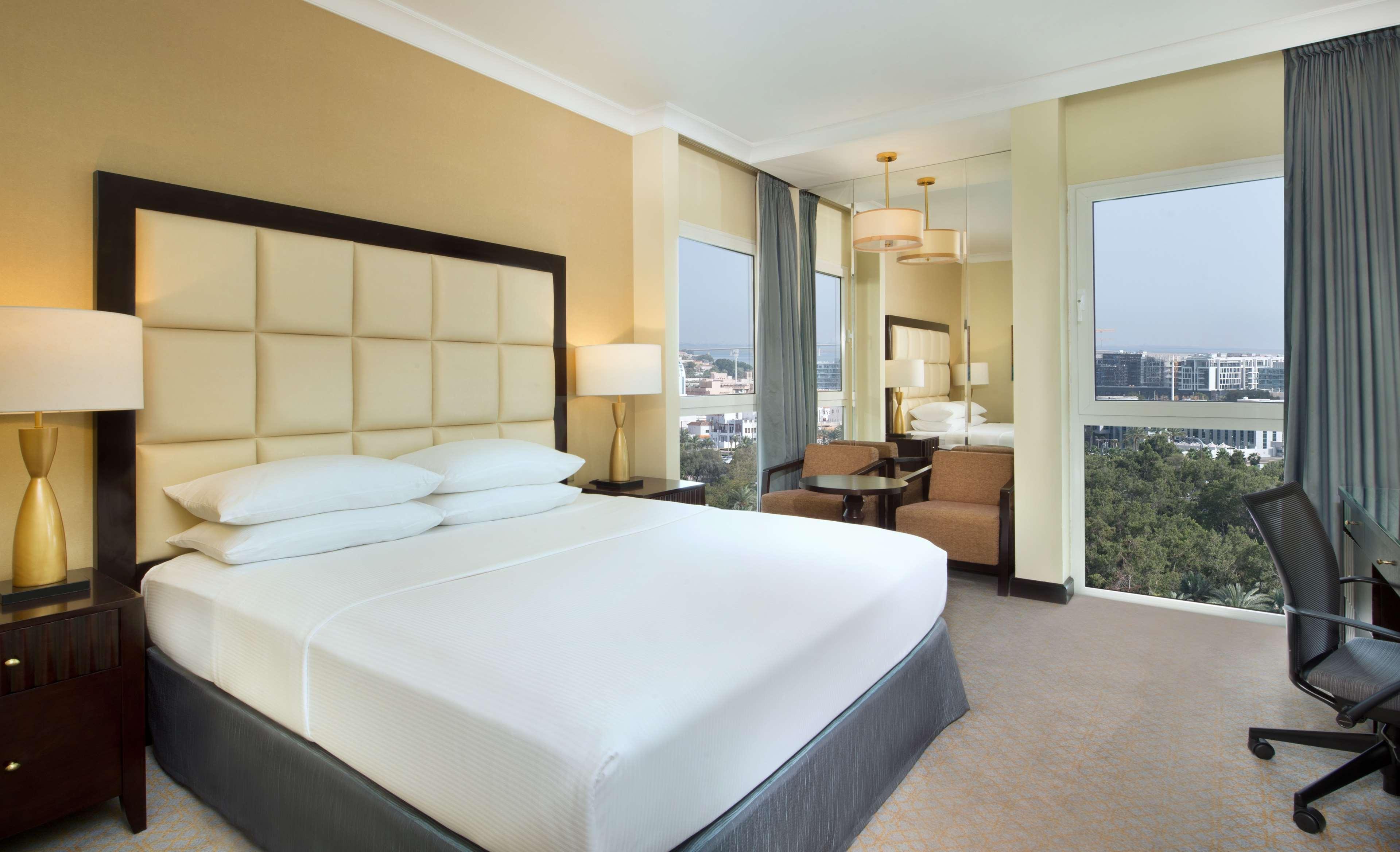 Radisson Blu Hotel & Resort, Abu Dhabi Corniche Extérieur photo The photo shows a modern hotel room featuring a large bed with white linens and a gray bed skirt. The headboard is upholstered in a light-colored fabric. There are bedside lamps on either side of the bed. In the background, there are windows offering