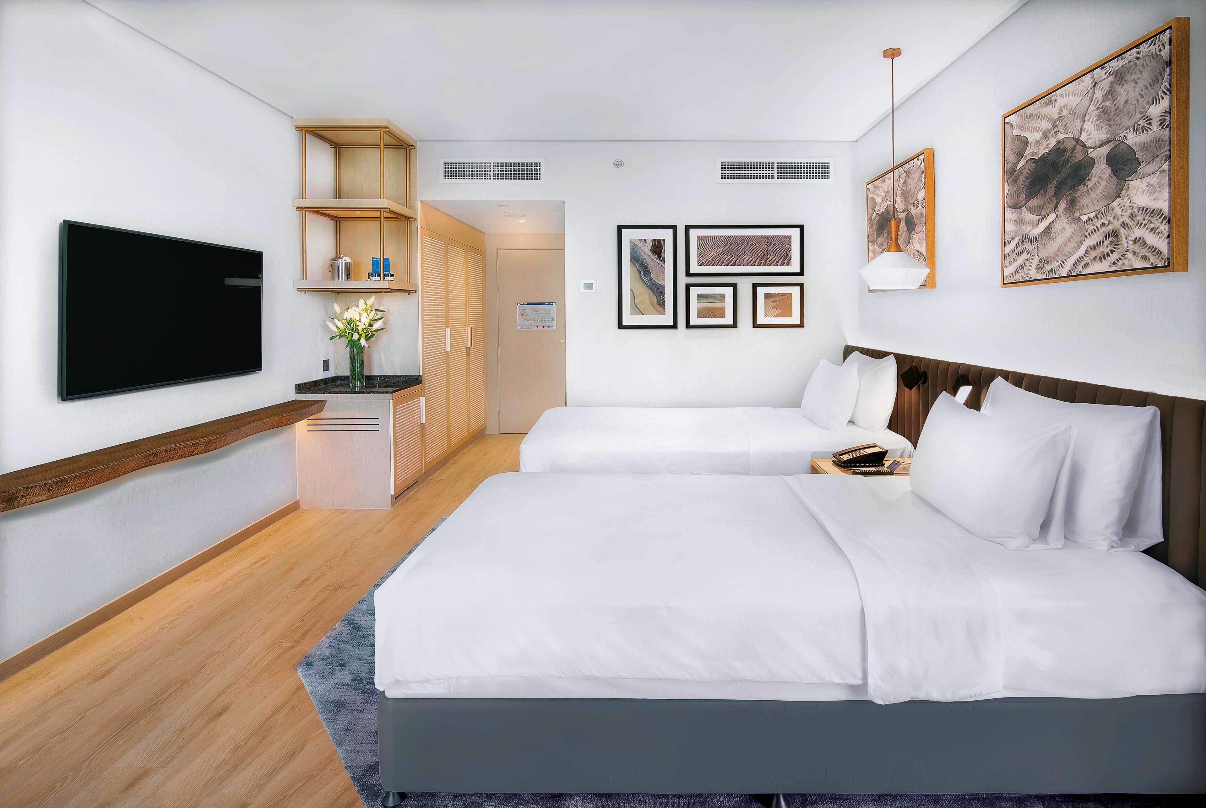 Radisson Blu Hotel & Resort, Abu Dhabi Corniche Extérieur photo The photo shows a modern hotel room featuring two beds with white bedding. There's a flat-screen TV mounted on the wall opposite the beds. The room has light-colored wooden flooring and a large window, allowing natural light to enter. A small kitchen