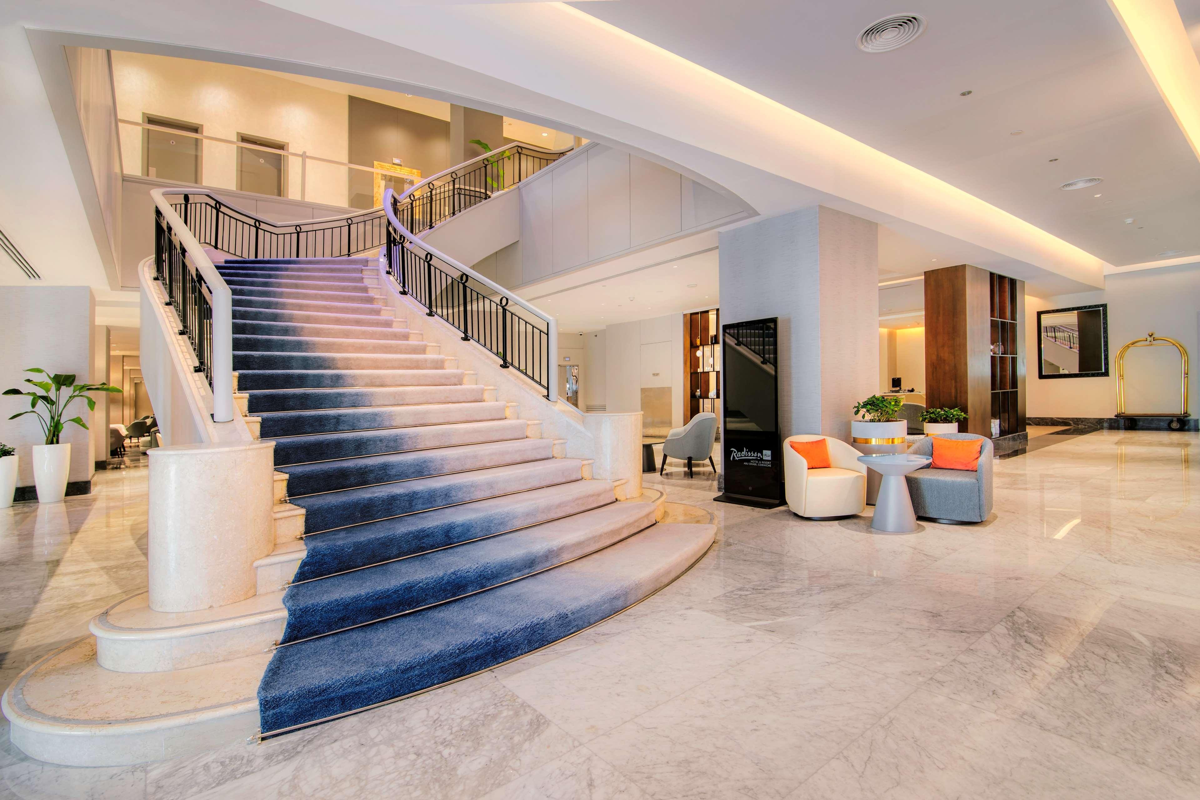 Radisson Blu Hotel & Resort, Abu Dhabi Corniche Extérieur photo The photo shows a welcoming hotel lobby with a prominent staircase. The staircase features a stylish blue carpet that gradually transitions in color. The lobby is bright and spacious, with modern decor. To one side, there are seating arrangements con