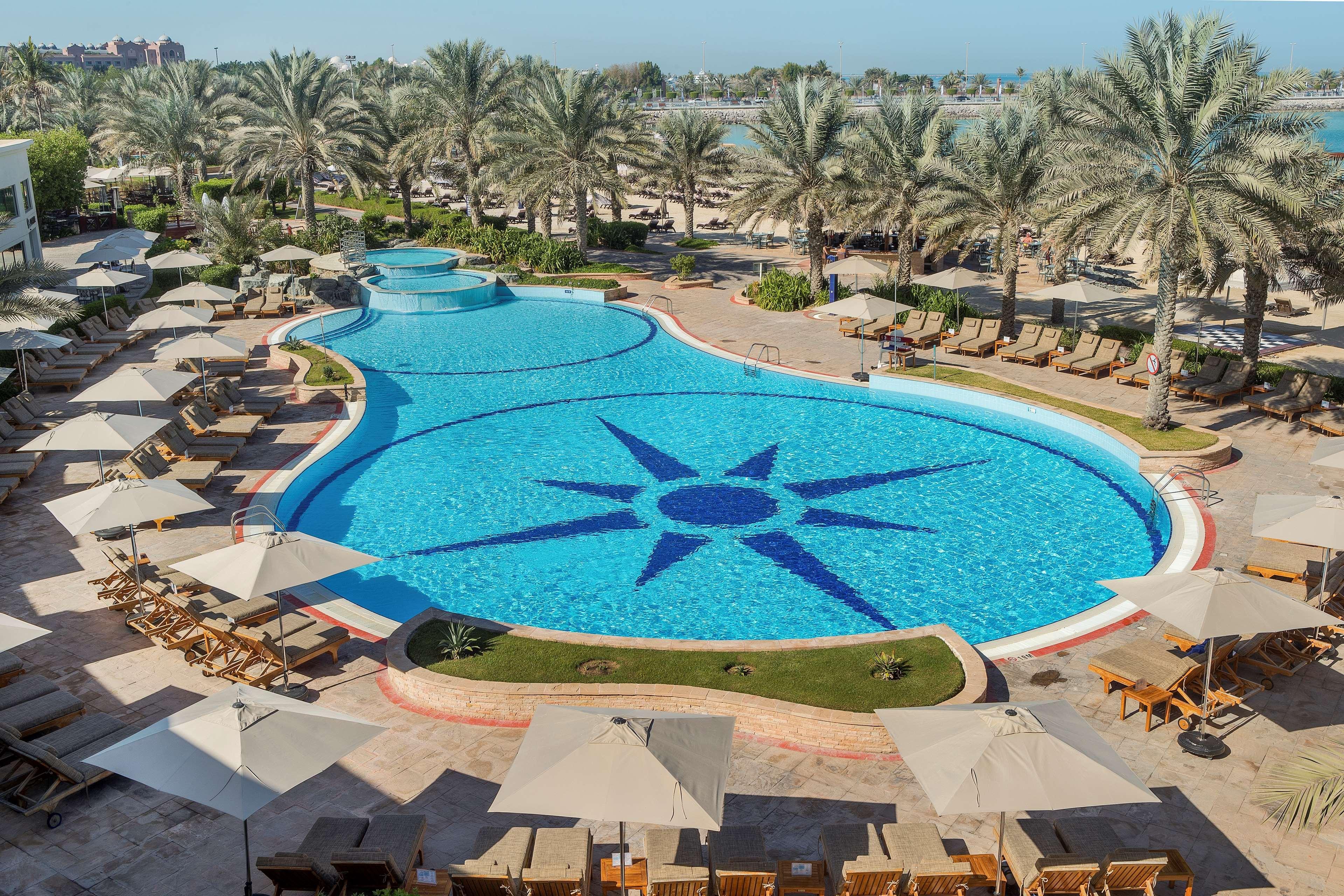 Radisson Blu Hotel & Resort, Abu Dhabi Corniche Extérieur photo The image depicts a large, beautifully designed swimming pool shaped like a freeform design, featuring a prominent star pattern at its center. Surrounding the pool are numerous lounge chairs with light-colored cushions and umbrellas, providing shade.