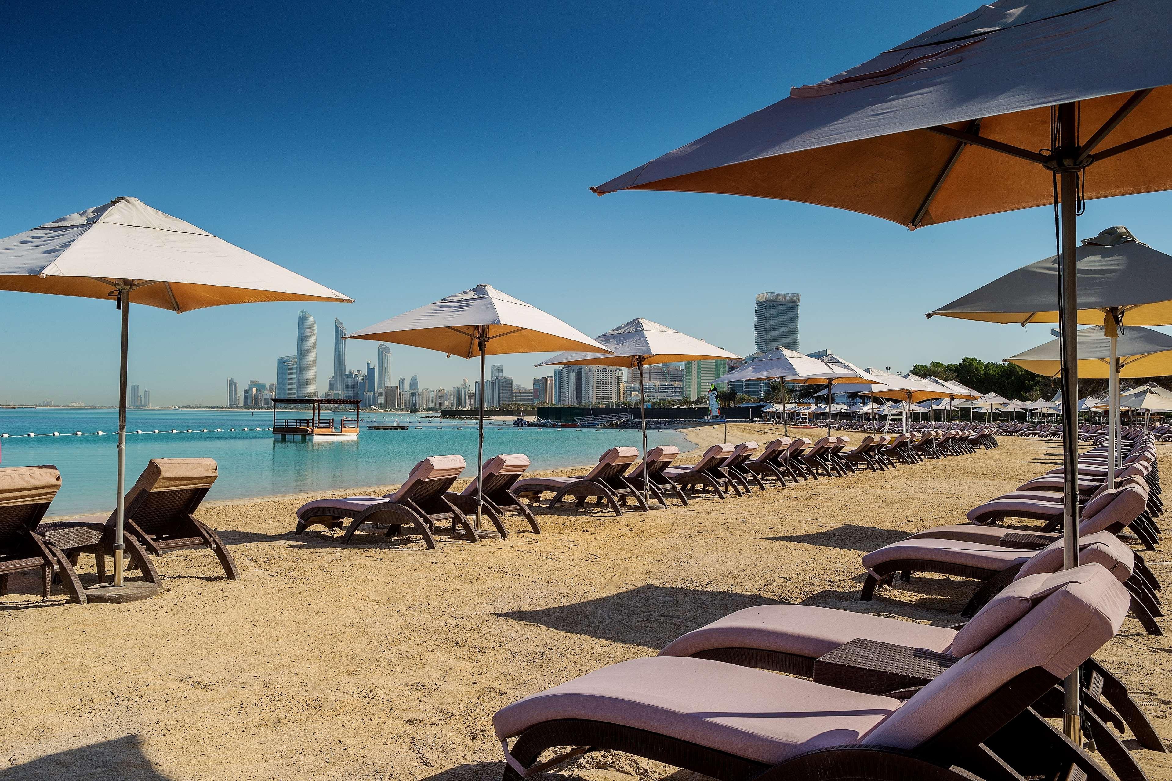 Radisson Blu Hotel & Resort, Abu Dhabi Corniche Extérieur photo The photo depicts a serene beach scene featuring rows of lounge chairs and umbrellas arranged along the sandy shore. In the background, there are modern skyscrapers visible, suggesting a developed coastal city. The water is calm and turquoise, adding