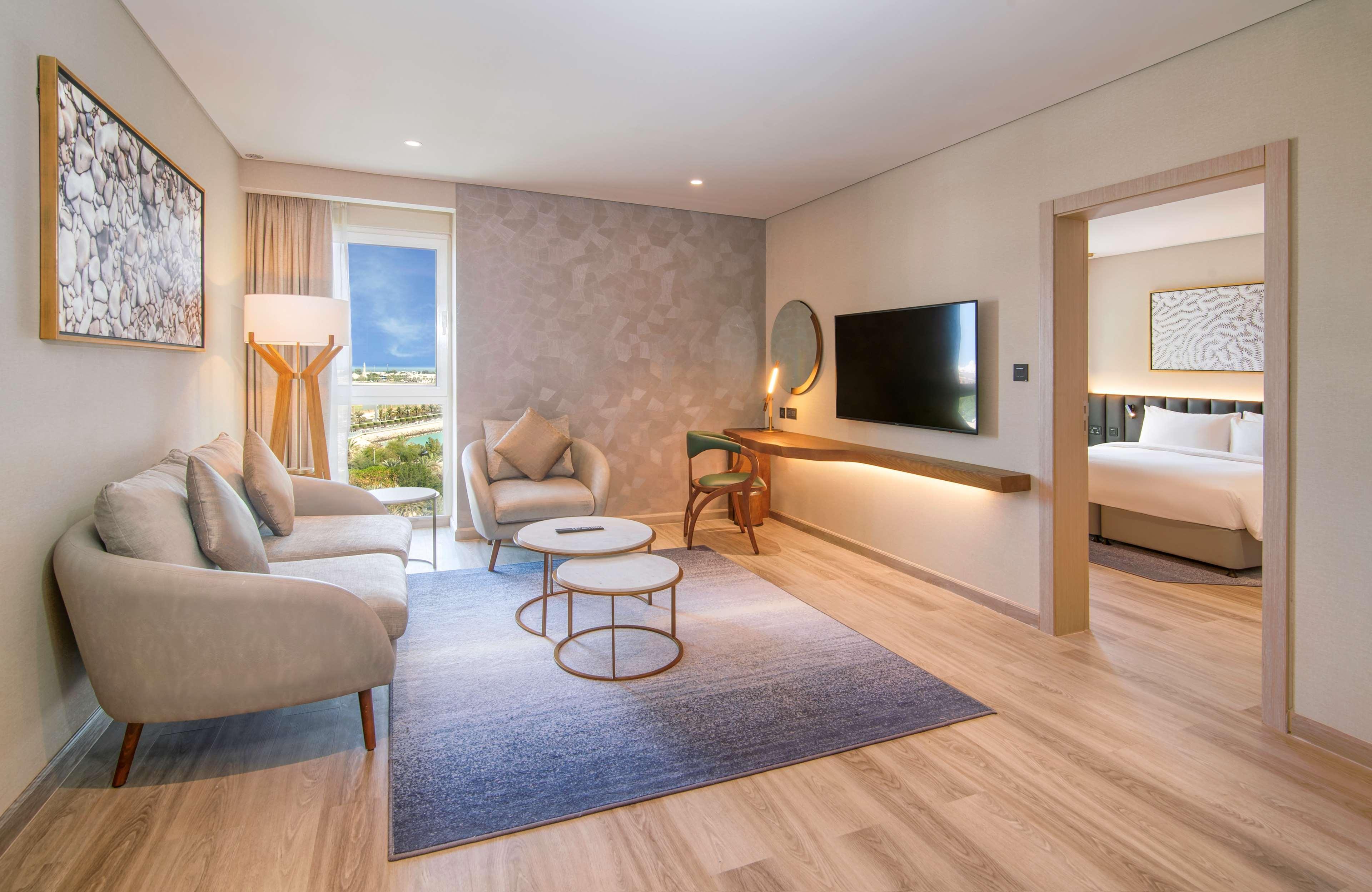 Radisson Blu Hotel & Resort, Abu Dhabi Corniche Extérieur photo The photo shows a modern hotel room featuring a living area. It includes two comfortable light-colored armchairs and a small round coffee table in front. The room has a large window with curtains that allow natural light to enter, and beyond the wind