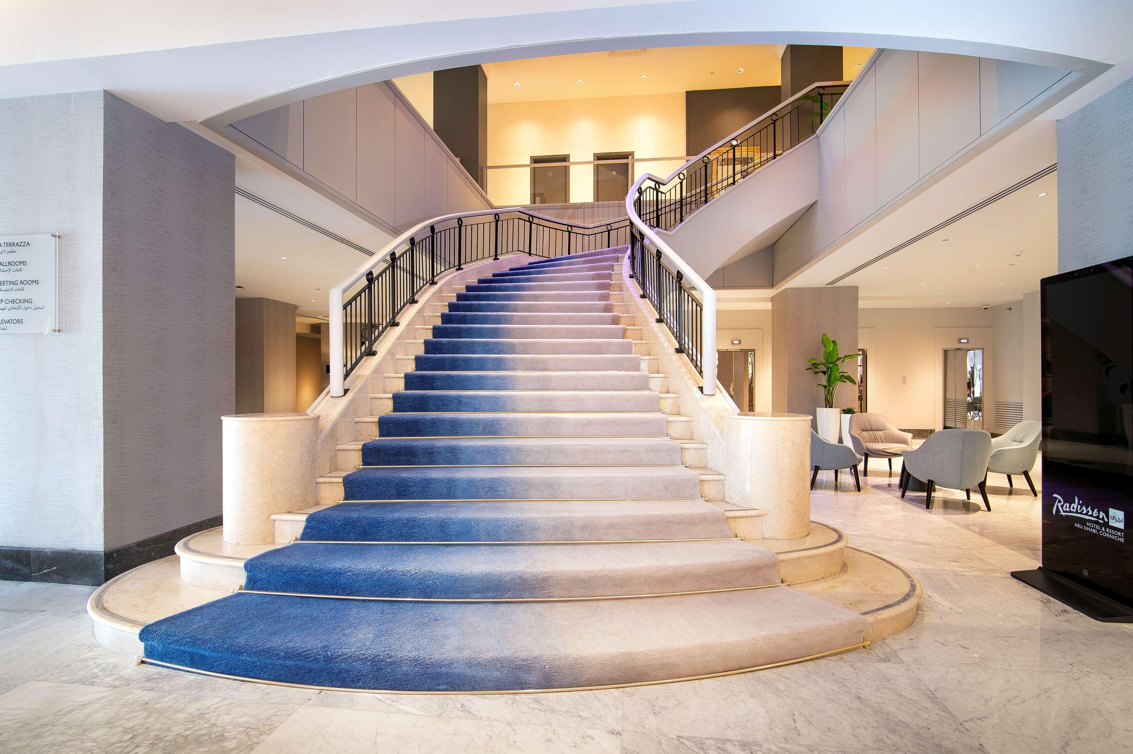 Radisson Blu Hotel & Resort, Abu Dhabi Corniche Extérieur photo The photo depicts an elegant staircase inside a building, likely a hotel or a similar venue. The staircase is wide with a gentle curve, featuring a blue carpet that transitions in color from light to dark as it ascends. The walls surrounding the stai