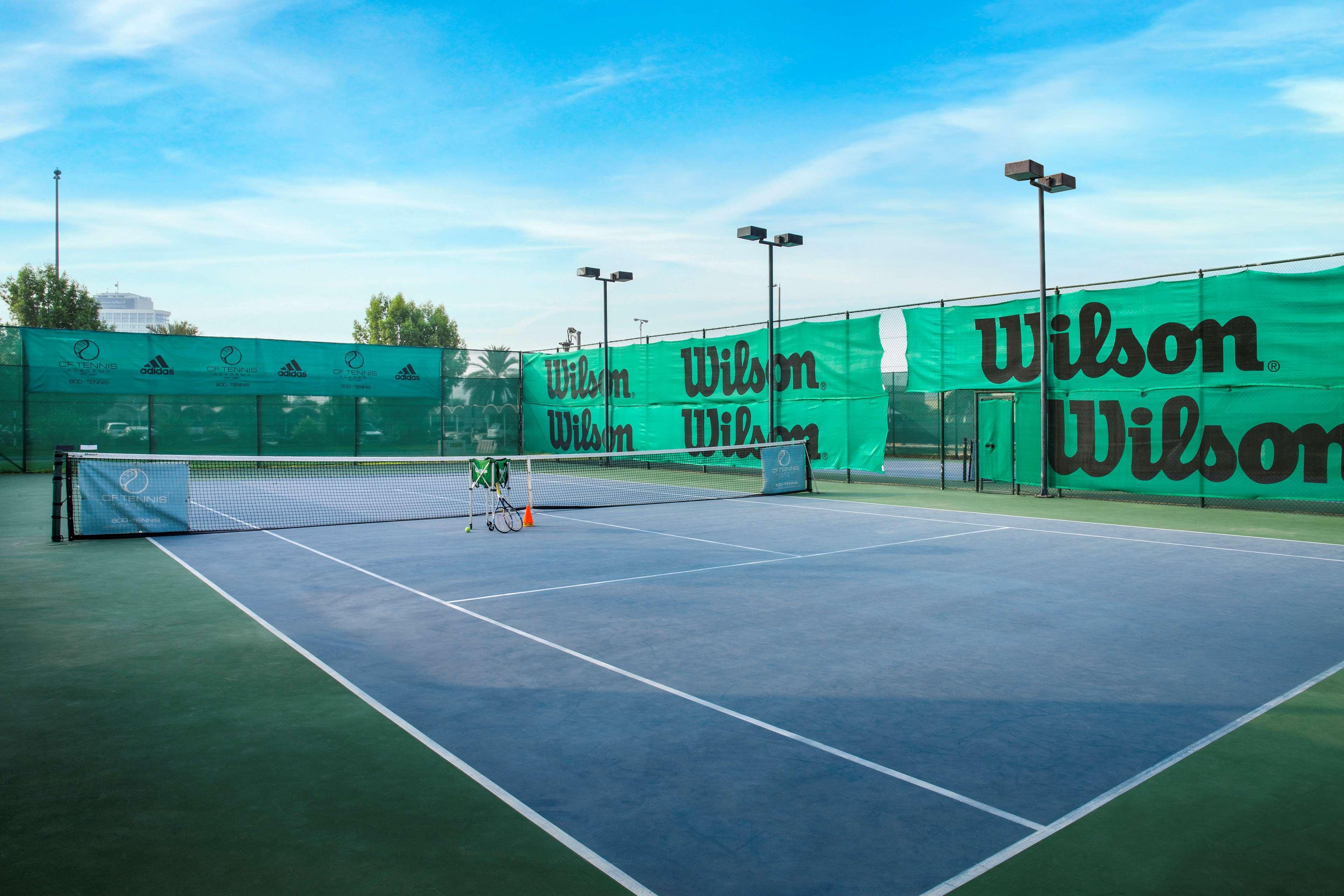 Radisson Blu Hotel & Resort, Abu Dhabi Corniche Extérieur photo The photo shows a view of a tennis court surrounded by green nets and fencing featuring the brand name "Wilson." The court itself has a hard surface with marked lines for play. There are several tennis equipment items, such as a few rackets and possi