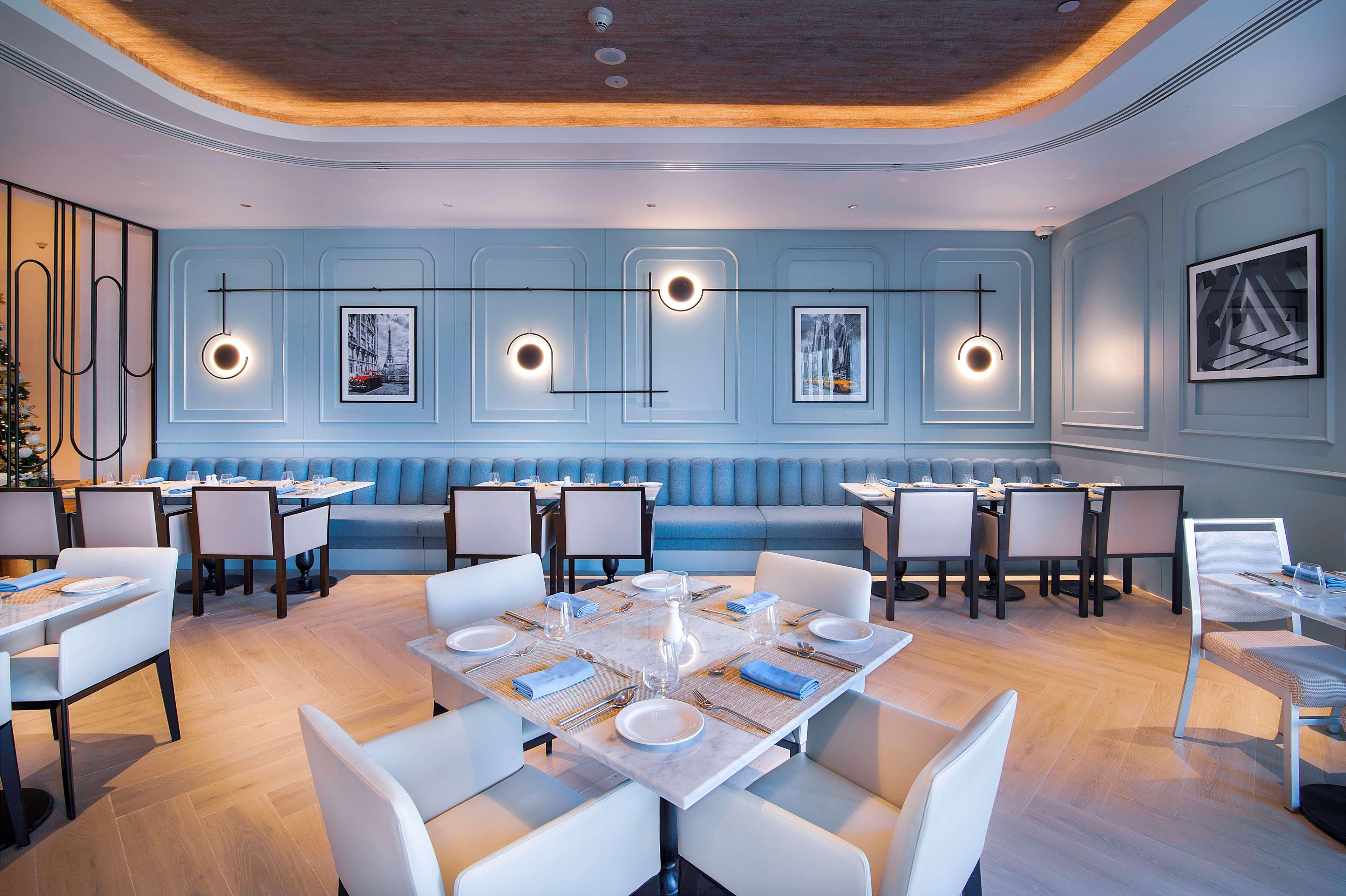 Radisson Blu Hotel & Resort, Abu Dhabi Corniche Extérieur photo The photo shows a modern restaurant interior. The walls are painted in a soft blue, complemented by a decorative paneling design. There are multiple circular light fixtures hanging from the ceiling, providing ambient lighting. The seating includes a 