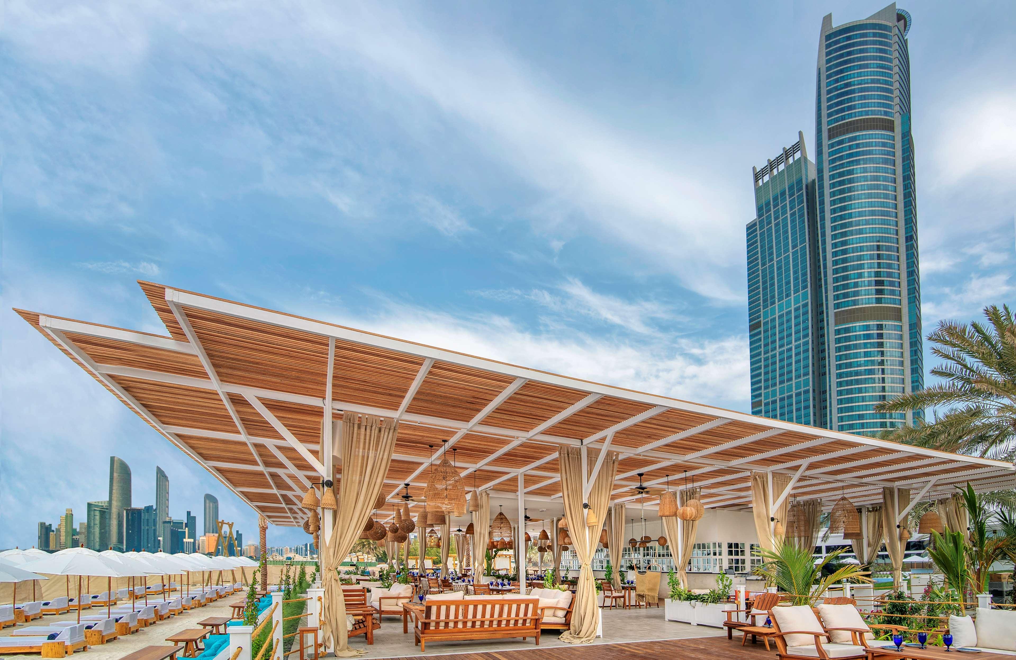 Radisson Blu Hotel & Resort, Abu Dhabi Corniche Extérieur photo The photo shows a modern outdoor lounge or café area situated by a body of water, likely a beach or waterfront. The structure has a large, open roof with wooden beams and curtains providing shade. There are comfortable seating arrangements, such as s