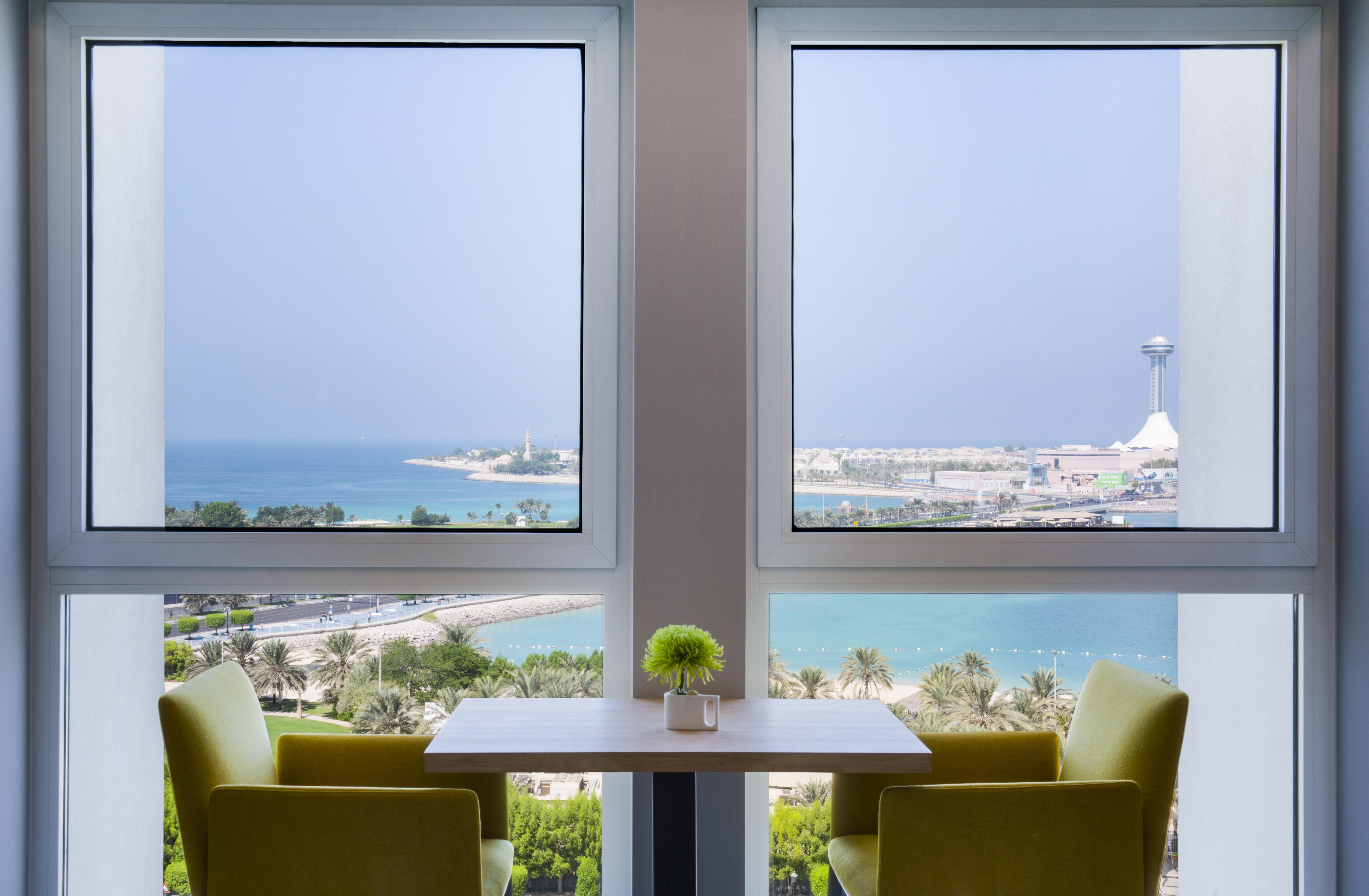 Radisson Blu Hotel & Resort, Abu Dhabi Corniche Extérieur photo The photo shows a serene interior space featuring two large windows that frame a beautiful view of the ocean and coastline. The scene is bright, with sunlight filtering in, and there are two green chairs positioned around a small table. A small plant