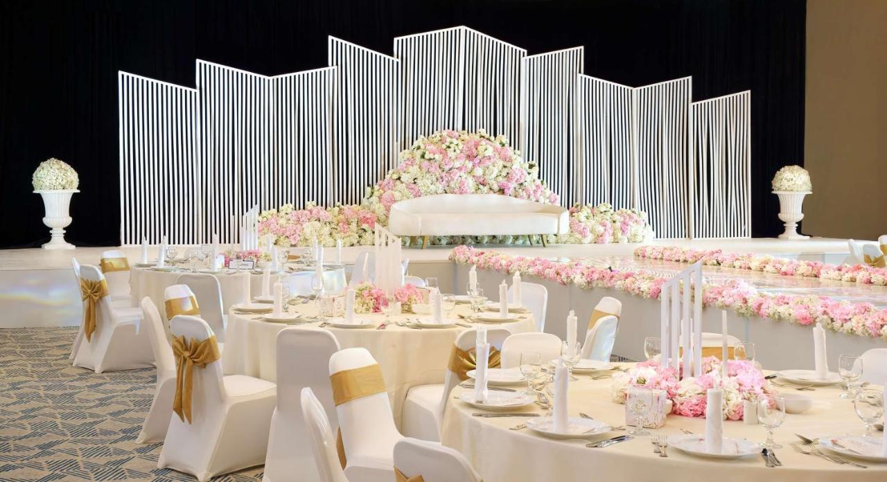 Radisson Blu Hotel & Resort, Abu Dhabi Corniche Extérieur photo The photo features an elegantly decorated banquet hall set up for a special event, likely a wedding. In the background, there is a large backdrop adorned with vertical white lines and a lush arrangement of pink flowers. The seating area consists of r