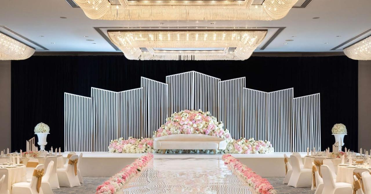 Radisson Blu Hotel & Resort, Abu Dhabi Corniche Extérieur photo The photo shows a beautifully decorated wedding venue. In the center, there's a large, elegant white sofa on a stage, surrounded by a backdrop consisting of vertical white lines that create a modern, geometric design. In front of the sofa, a lush arr